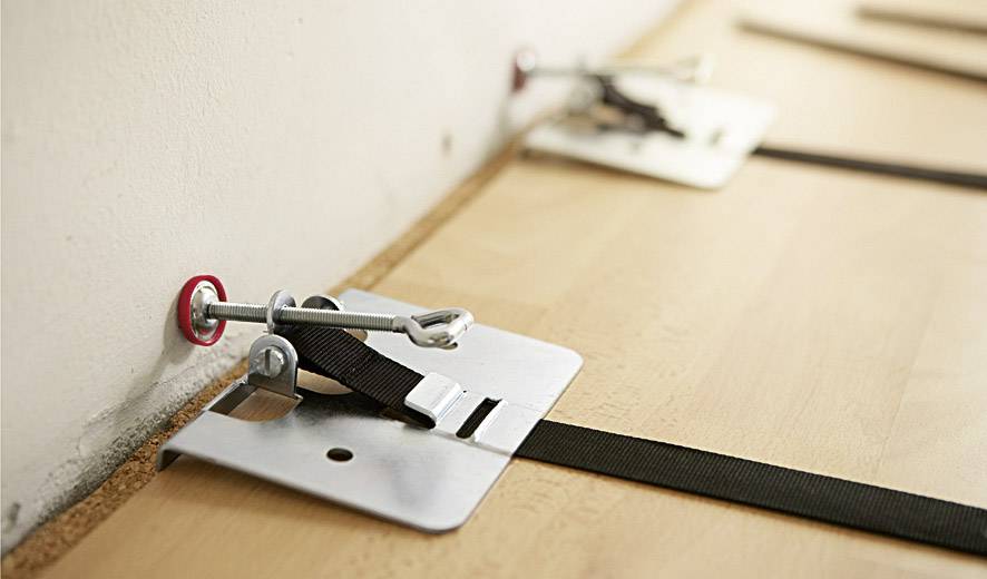 Bodenbelag wird mithilfe von Spannvorrichtungen straff an der Wand fixiert. Holzboden und Spanngurte sind sichtbar.