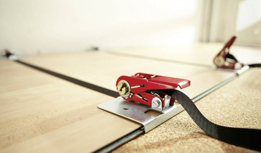 Ein Spanngurt ist an einer Holzplatte befestigt, um diese sicher zu fixieren. Die Szene zeigt eine Nahaufnahme der Befestigung.