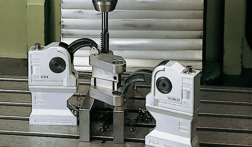 Eine Maschine in einer Werkstatt, die ein Werkstück mit zwei Klemmen hält. Ein Bohrer ist über dem Werkstück positioniert.