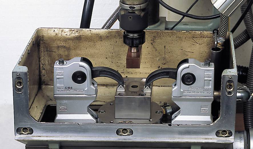 Eine industrielle Maschine führt einen Fertigungsprozess am Metallwerkstück durch, gehalten von zwei grauen Halterungen in der Mitte.