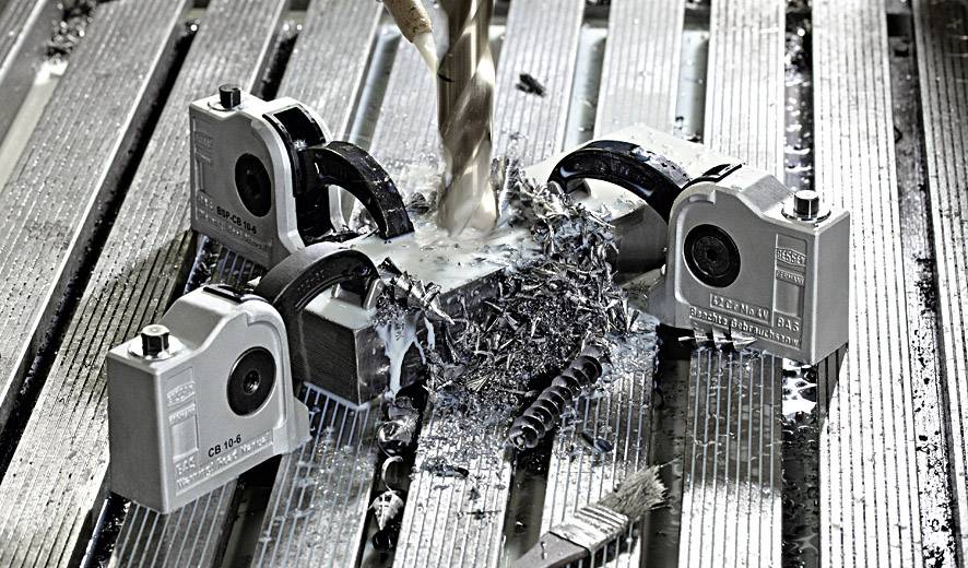 Bohrgerät bearbeitet Metallplatte mit Spänen umgeben. Mehrere Spannvorrichtungen halten das Werkstück fest.