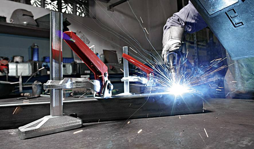 Ein Schweißer arbeitet in einer Werkstatt, Funken fliegen. Metallteile sind mit Klemmen befestigt, Arbeitsschutzkleidung wird getragen.