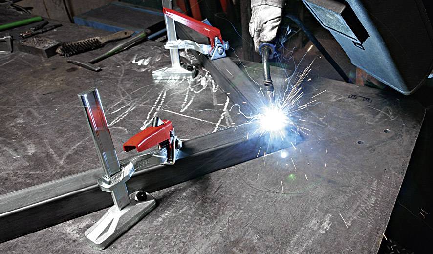 Eine Person schweißt Metallteile auf einem Tisch, umgeben von Klammern und Funkenschlag. Schutzkleidung wird getragen.