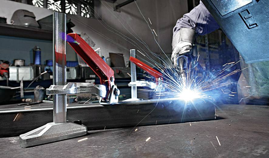 Eine Person schweißt an einem Metallstück, während Funken sprühen. Im Hintergrund sind Werkzeuge und Werkbänke zu sehen.