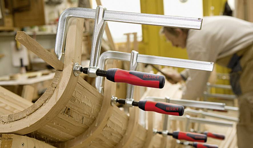 Holzbearbeitung in Werkstatt: Mehrere Schraubzwingen halten gebogene Holzteile. Handwerker im Hintergrund arbeitet konzentriert.