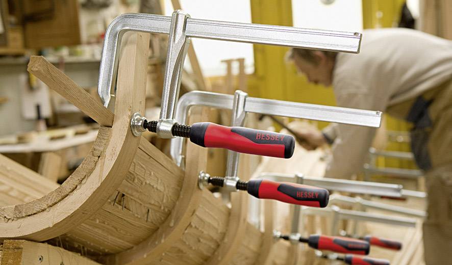 Eine Person arbeitet in einer Holzwerkstatt. Mehrere Schraubzwingen halten Holzstücke zusammen. Der Fokus liegt auf der Holzbearbeitung.
