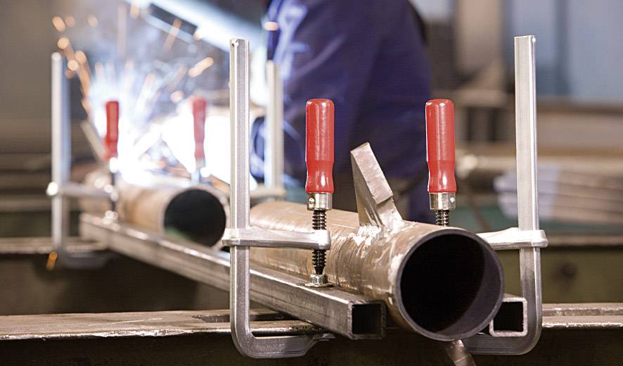 Ein Arbeiter schweißt Metallrohre, die mit Klammern gesichert sind. Funken fliegen, was auf einen aktiven Schweißvorgang hinweist.