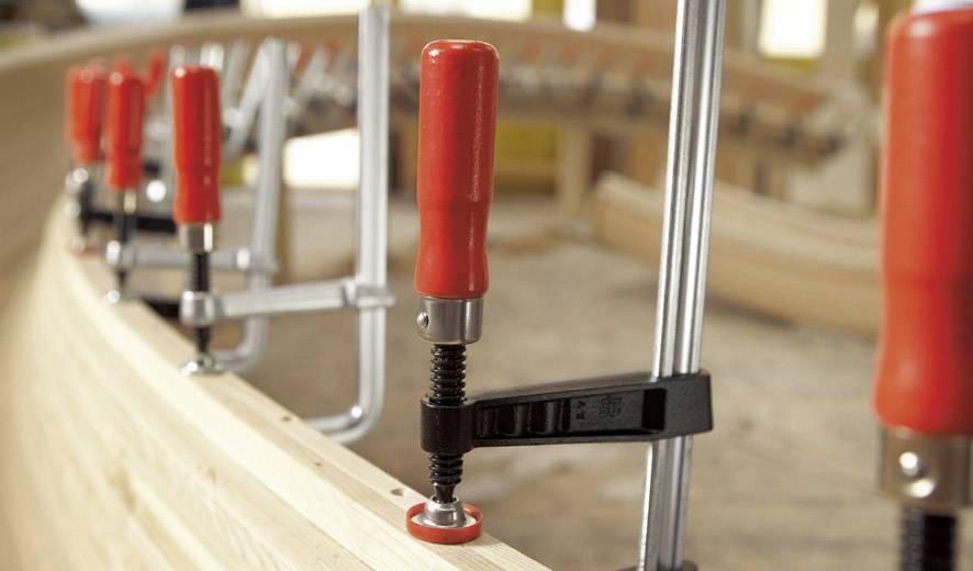 Mehrere Klemmen mit roten Griffen halten Holzplanken in einer Werkstatt fixiert.