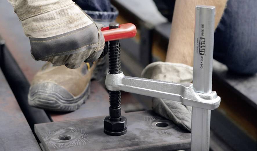 Eine Person mit Handschuhen benutzt eine rote Schraubzwinge, um ein Werkstück zu fixieren. Das Bild zeigt handwerkliche Arbeit.