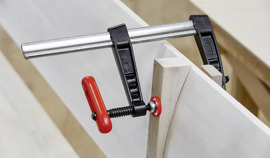 Metallzwinge hält zwei Holzteile zusammen. Eine rote Schraube dient zum Anziehen. Werkstattumgebung sichtbar im Hintergrund.