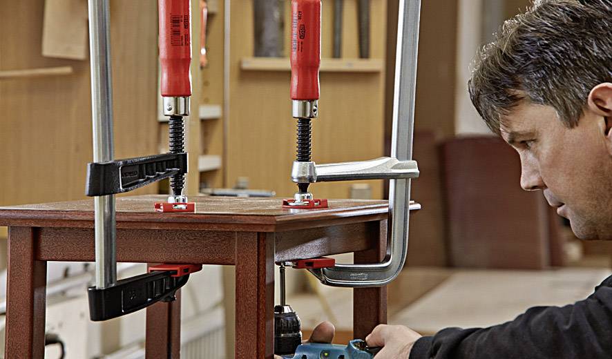 Ein Handwerker befestigt mit Schraubzwingen zwei Holzteile an einem Tisch und arbeitet sorgfältig mit einem blauen Werkzeug daran.