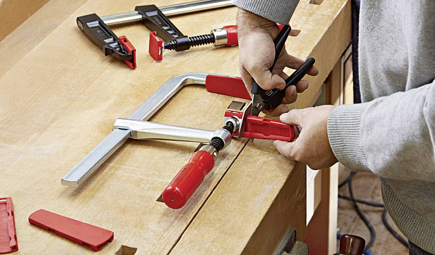 Eine Person benutzt eine Zwinge auf einem Holztisch. Umherliegende rote Klammern und Werkzeuge deuten auf eine Arbeitssituation hin.