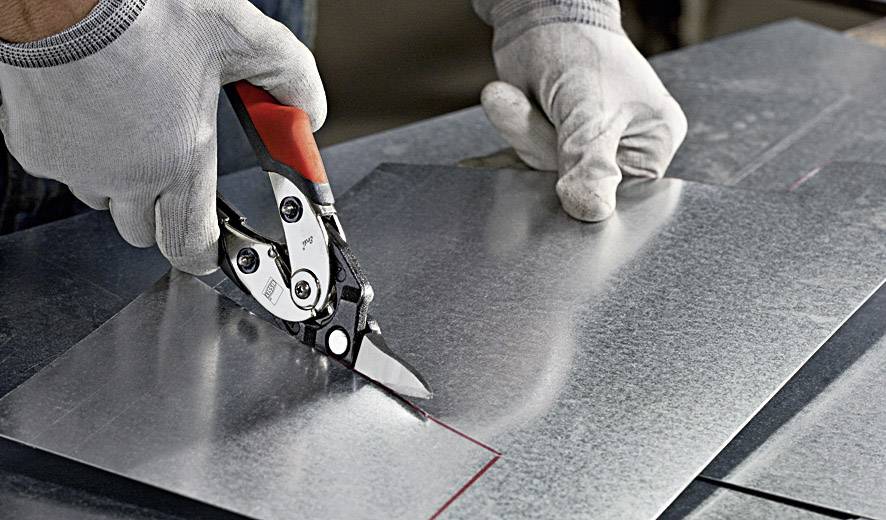 Person in grauen Handschuhen schneidet Blech mit einer Metallschere.