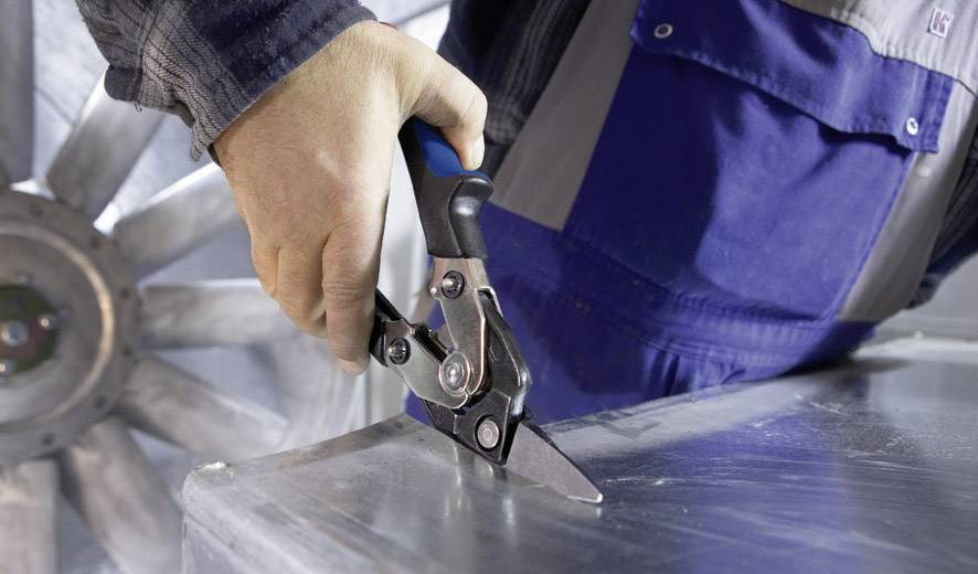 Eine Hand hält eine Metallschere und schneidet Blech. Im Hintergrund sind industrielle Details sichtbar.