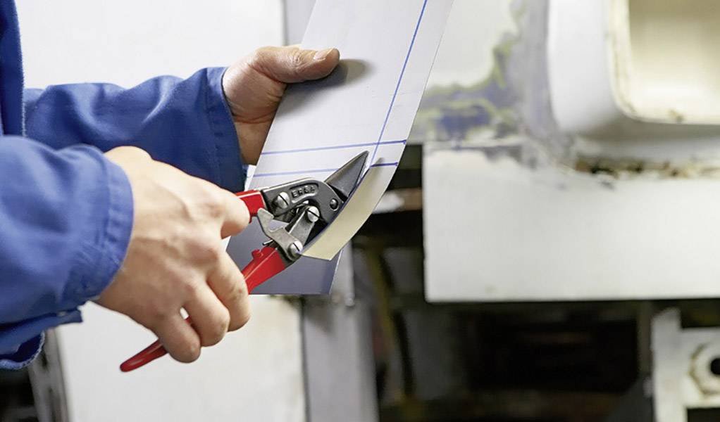 Eine Person in blauer Arbeitskleidung schneidet ein Blech mit einer Metallschere. Im Hintergrund sind Werkstattdetails sichtbar.