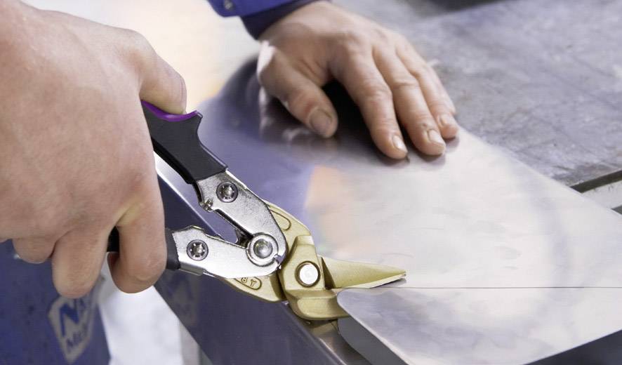 Eine Person schneidet mit einer Metallschere durch ein Blech. Die linke Hand hält die Schere, während die rechte Hand das Blech stützt.