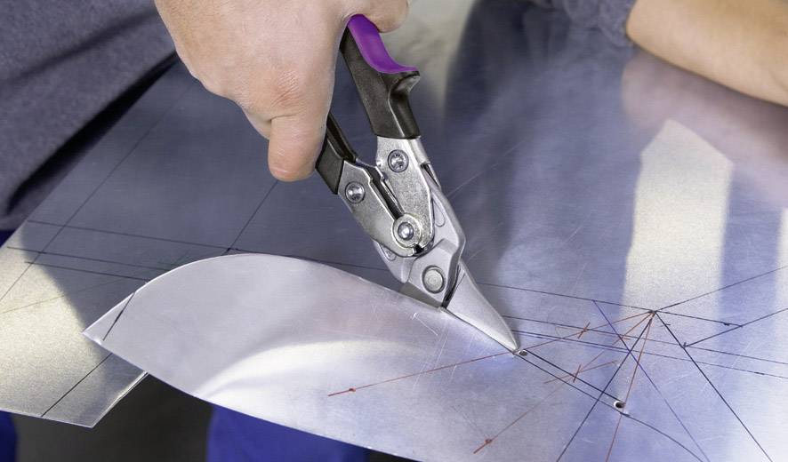 Eine Person schneidet ein Blech mit einer Metallschere, sichtbare Markierungen auf dem Metall zeigen die Schnittlinien.