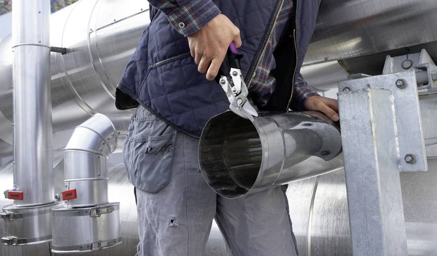 Eine Person schneidet mit einer Metallschere ein rundes Metallrohr neben industriellen Anlagen. Person trägt Arbeitskleidung.