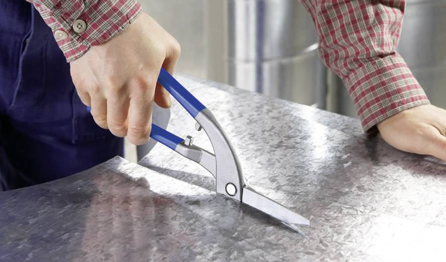 Eine Person schneidet mit einer blauen Metallschere ein Stück Blech in einer Werkstatt.