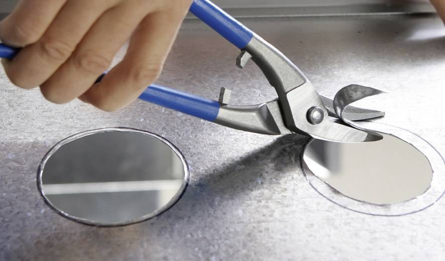 Eine Hand hält eine Metallschere und schneidet ein rundes Loch in ein Metallblech.