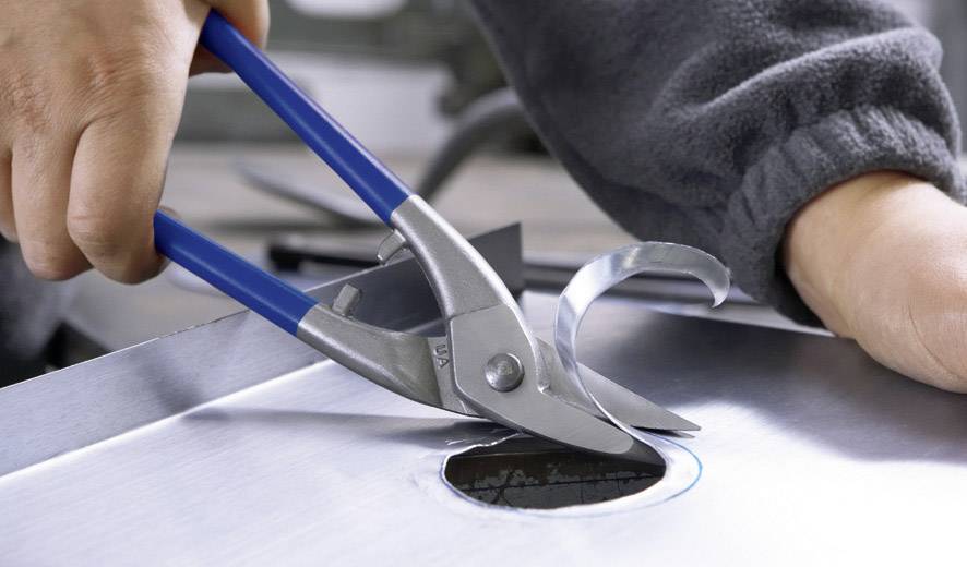 Eine Hand benutzt eine Blauschneiderzange, um ein rundes Loch in einem Metallblech zu schneiden. Das Werkzeug biegt Metallstücke auf.