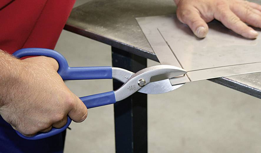 Person schneidet mit einer blauen Blechschere ein dünnes Metallblech auf einem Tisch.