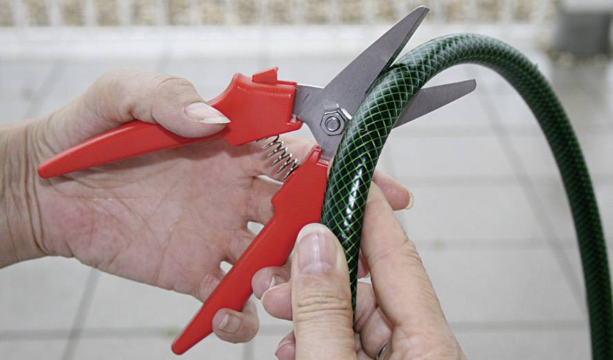Eine Person schneidet mit einer roten Schere einen grünen Gartenschlauch. Hände halten die Schere und den Schlauch fest.