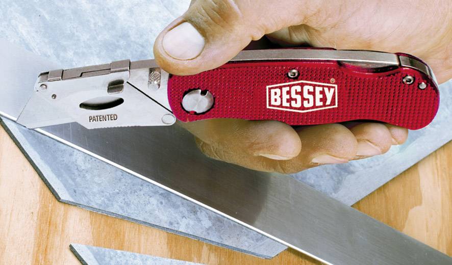 Eine Hand hält ein rotes Klappmesser mit der Aufschrift 'Bessey'. Trapezklinge und Metallteile liegen auf hölzernem Untergrund.