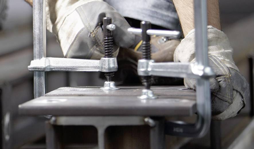 Eine Person mit Arbeitshandschuhen befestigt ein Metallstück mit Schraubzwingen an einem Tisch in einer Werkstatt.