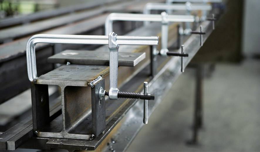 Mehrere Schraubzwingen halten Metallträger auf einer Werkbank. Sie sichern die Träger für Arbeiten in einer Werkstattumgebung.