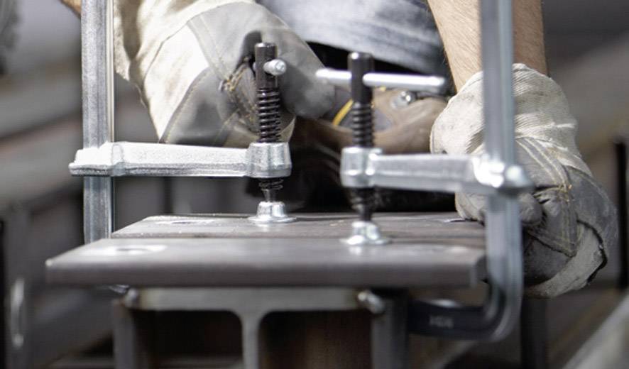 Ein Metallarbeiter justiert eine Schraubzwinge auf einem Metallstück. Hände tragen Arbeitshandschuhe. Fokus liegt auf Präzision und Technik.