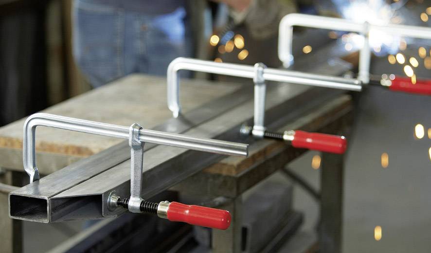 Nahaufnahme von Metallklemmen, die ein Metallstück auf einer Werkbank fixieren. Im Hintergrund sind Funken von Schweißarbeiten zu sehen.