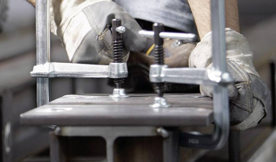 Nahaufnahme eines Metallarbeiters, der eine Klemme benutzt, um zwei Metallstücke zu fixieren. Arbeiter trägt Arbeitshandschuhe.