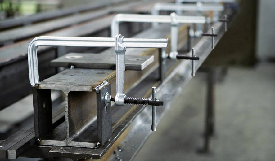 Ein Metallträger wird mit mehreren Metallzwingen fest auf einem Arbeitstisch fixiert. Die Umgebung wirkt industriell.