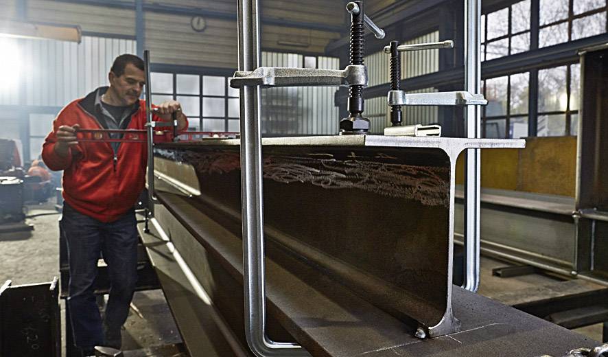 Ein Mann in roter Jacke justiert eine große Metallklemme in einer Werkstatt mit industrieller Ausstattung.