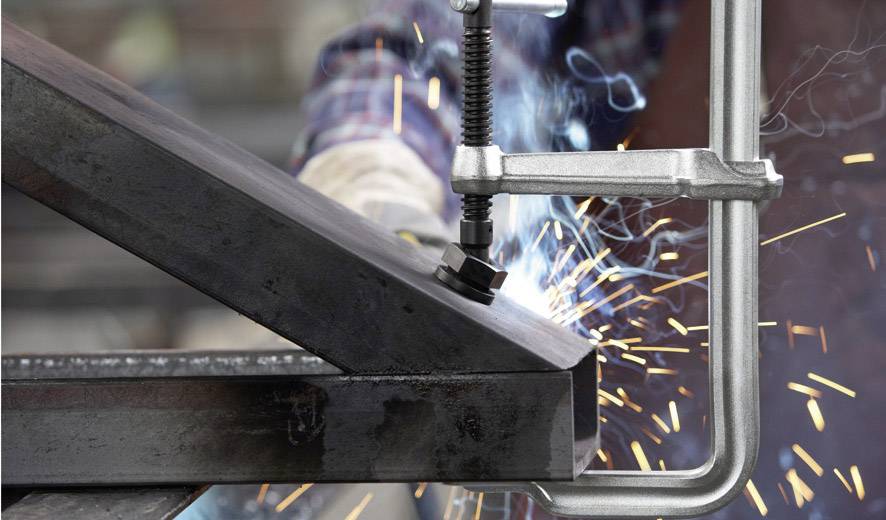 Eine Person in Arbeitskleidung schweißt zwei Metallstücke im rechten Winkel zusammen. Funken fliegen, während eine Klemme die Teile festhält.