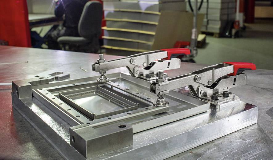 Eine Metallvorrichtung mit zwei Klemmen auf einem Arbeitstisch, von Schrauben stabilisiert. Eine Person arbeitet unscharf im Hintergrund.