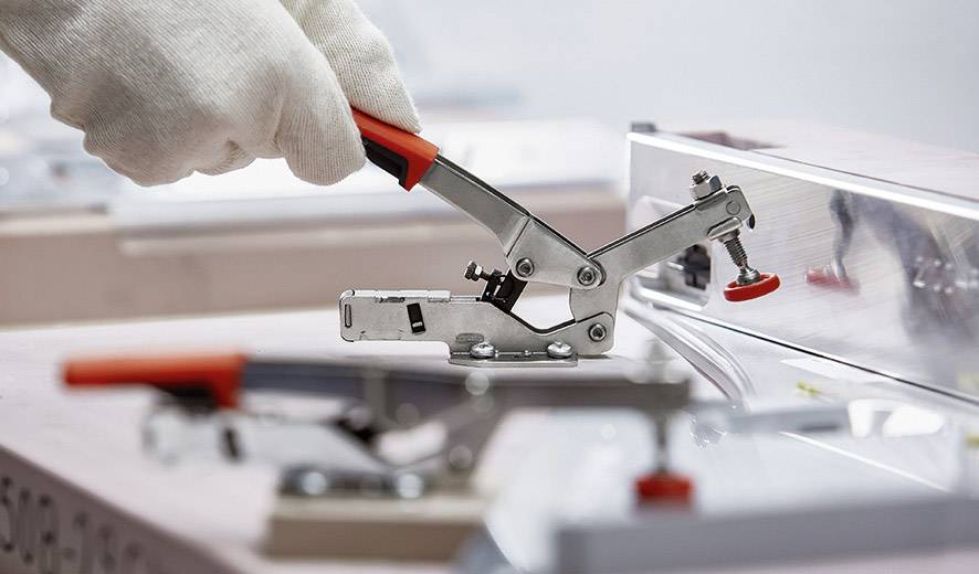 Eine behandschuhte Hand bedient ein Metallspannwerkzeug mit rotem Griff, das auf einer Maschine montiert ist.