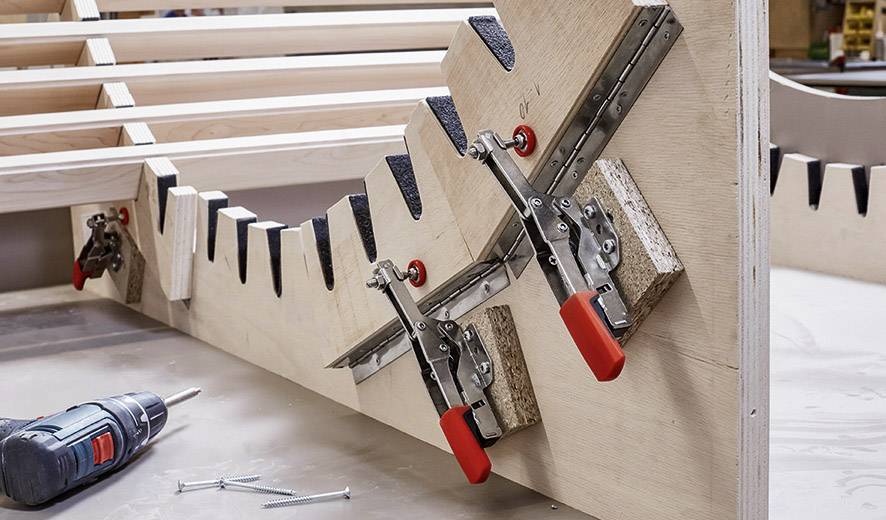 Holzwerkstück mit befestigten Klemmen während der Bearbeitung in einer Werkstatt. Eine Bohrmaschine und Schrauben liegen daneben.