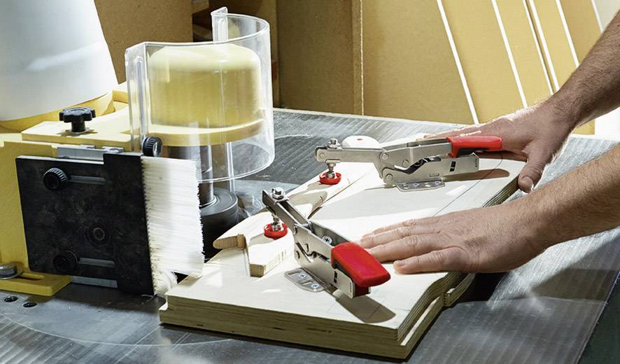 Maschine bearbeitet Holz, Hände halten das Brett, Sicherheitsvorrichtungen und Klemmen fixieren das Werkstück.