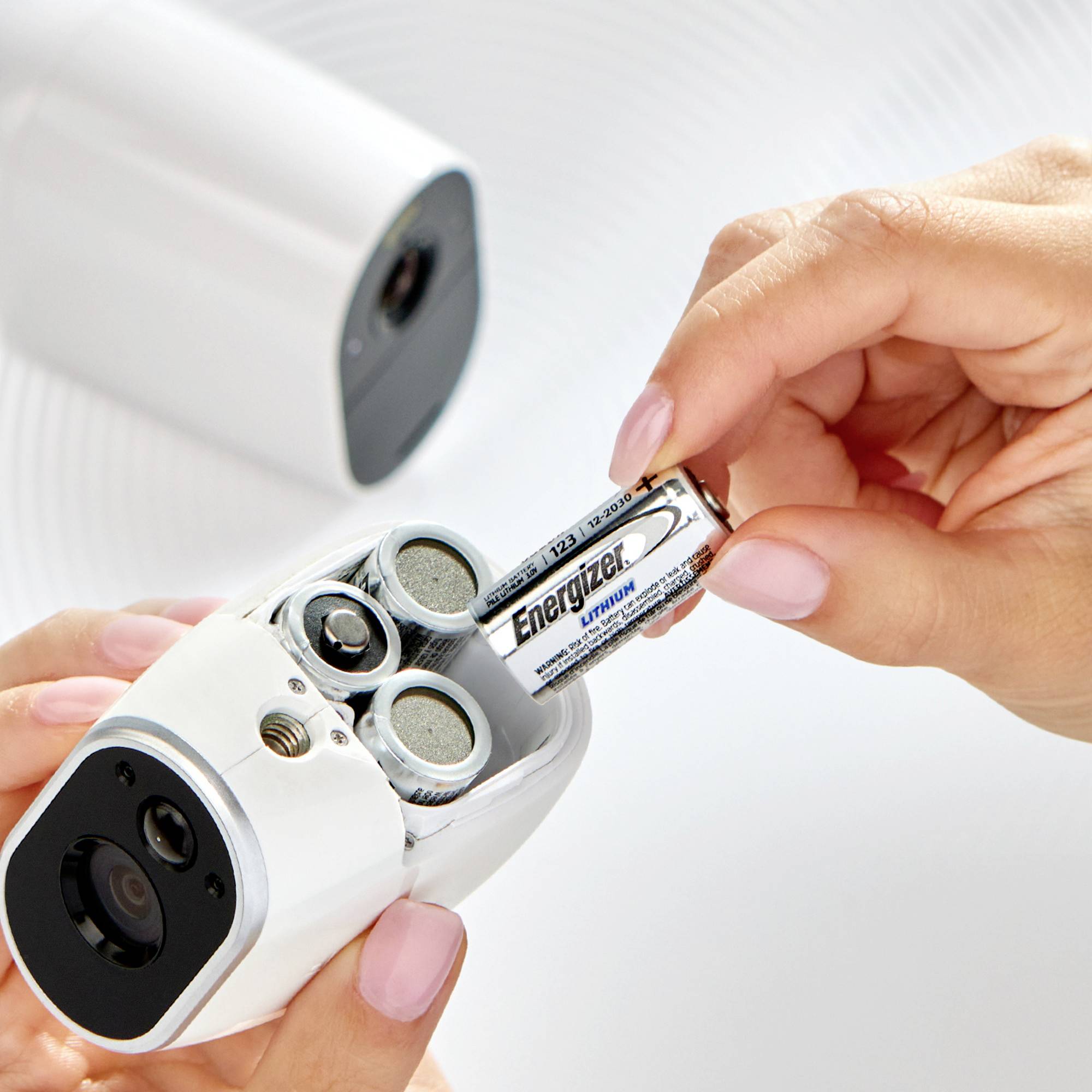 Eine Hand setzt eine Batterie in eine weiße, zylindrische Kamera ein, die auf einem Tisch liegt.