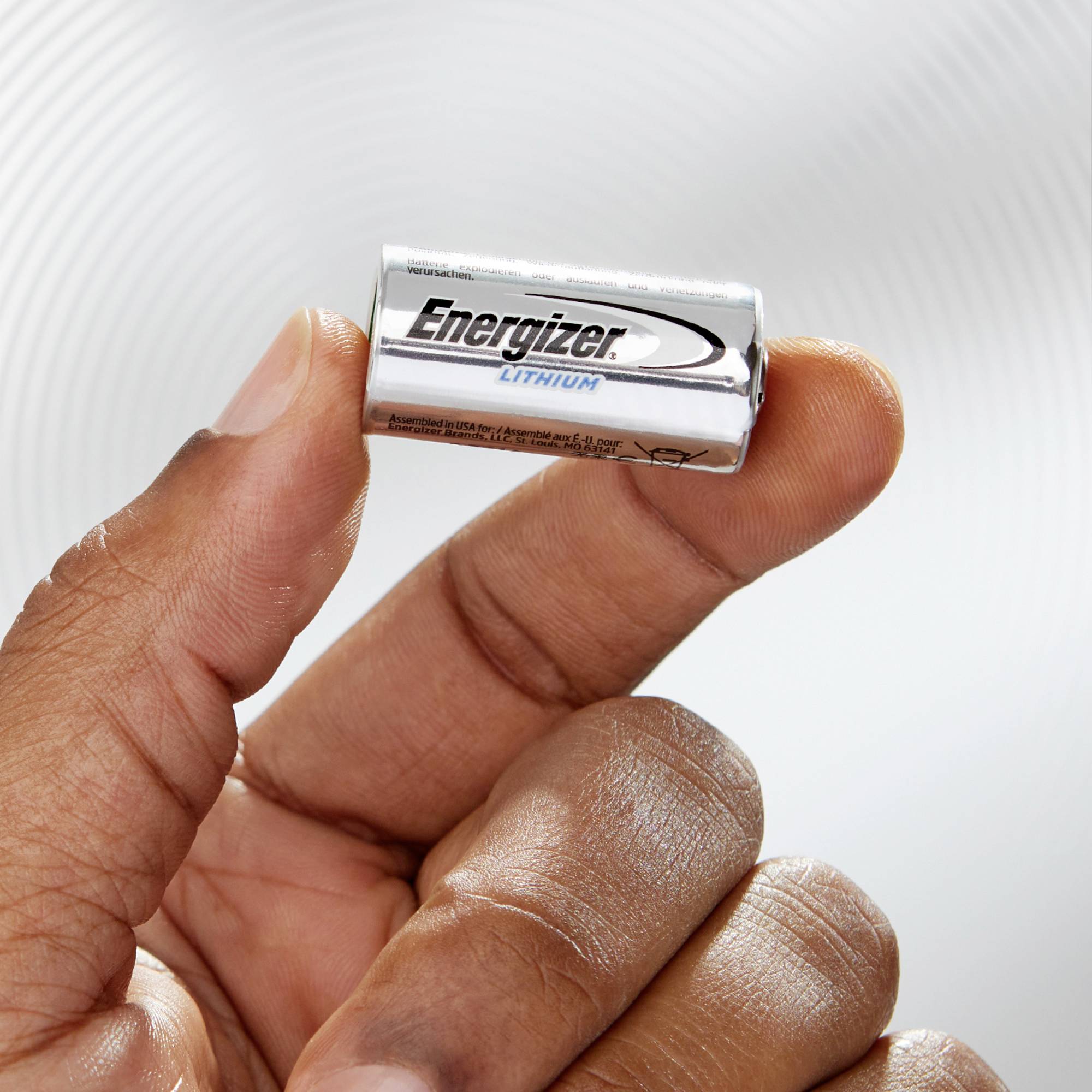 Eine Hand hält eine silberne 'Energizer'-Lithiumbatterie. Im Hintergrund unscharfe, helle Muster.