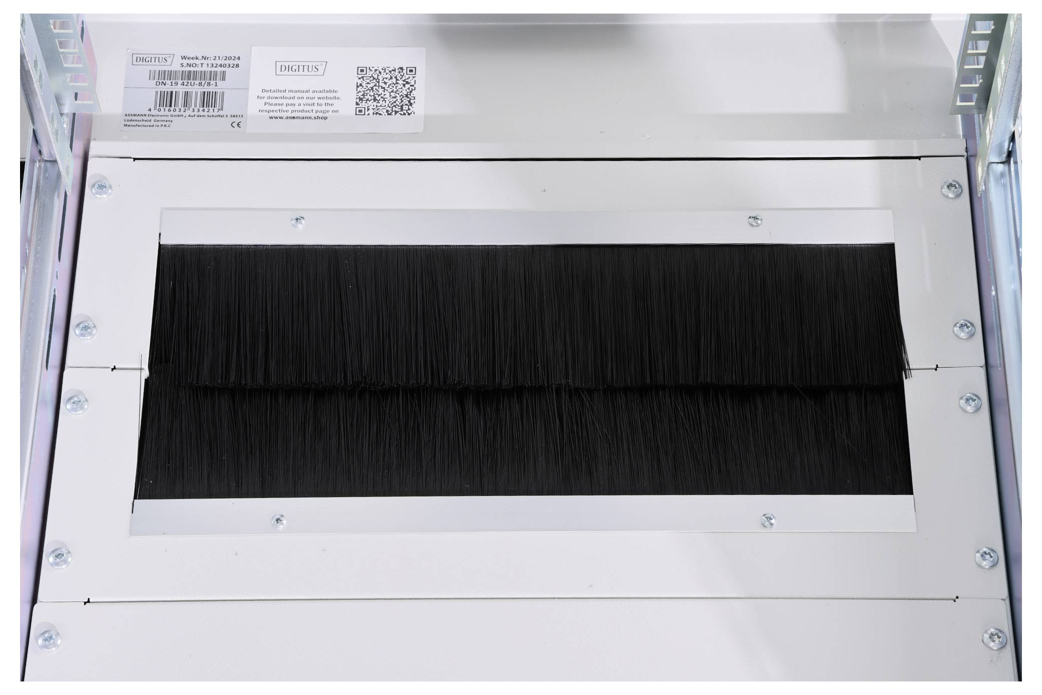 Ein metallener Kasten mit schwarzer Bürste im Inneren. Oben links sind ein Barcode und ein Etikett mit Detailinformationen sichtbar.