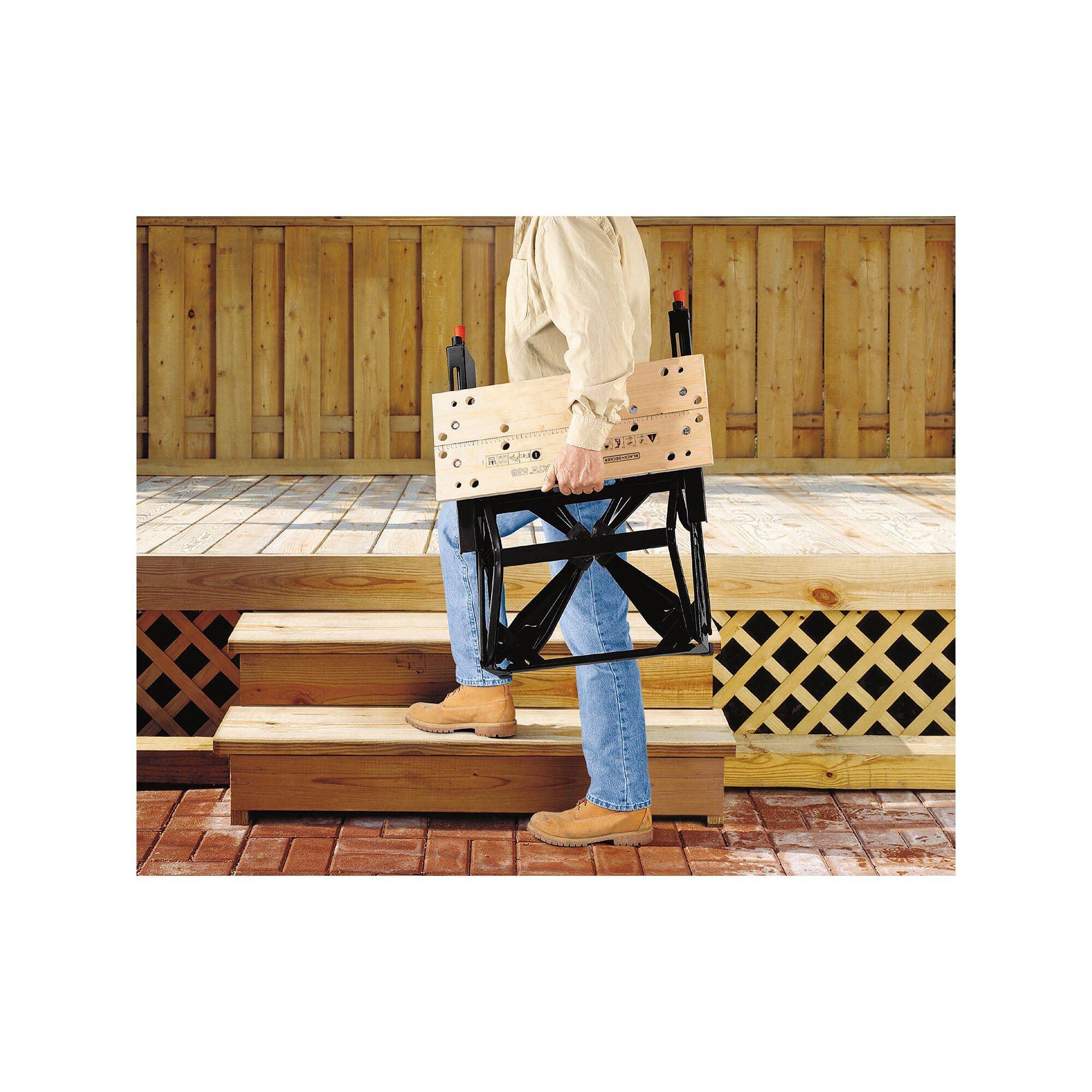 Eine Person trägt einen zusammengeklappten Klappstuhl aus Holz und Metall eine Holztreppe hinauf. Im Hintergrund ist eine hölzerne Veranda.