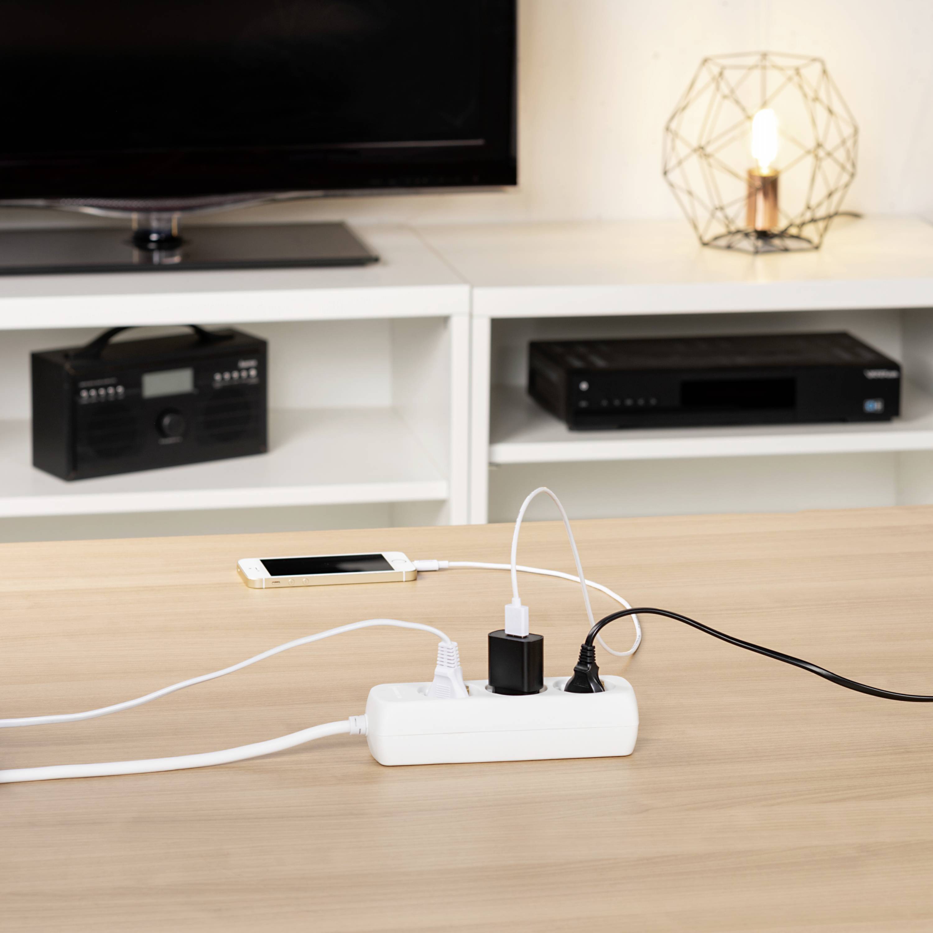 Ein Tisch mit Mehrfachsteckdose, in die mehrere Geräte wie Smartphone und Lampe eingesteckt sind. Im Hintergrund ein Fernseher und ein Radio.