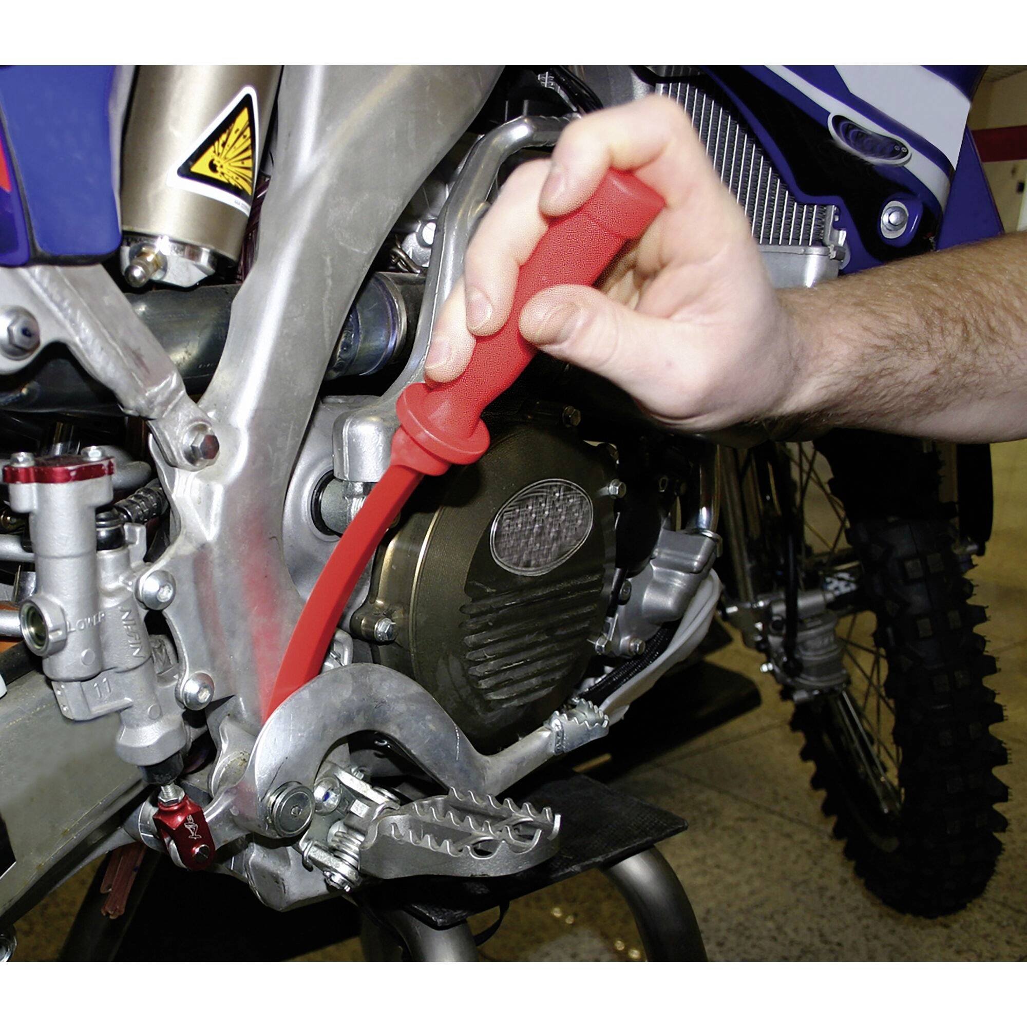 Person hält ein rotes Werkzeug und arbeitet an einem Motorrad. Fokus liegt auf Fußhebel und Kettenschloss des Motorrads.