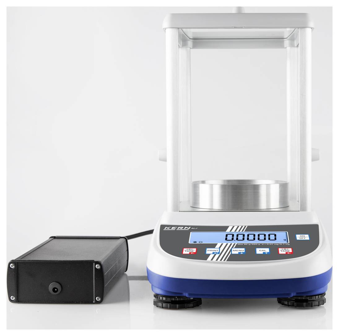 Eine digitale Laborwaage mit Schutzhaube, zeigt '0,0000' auf dem Display. Daneben befindet sich ein schwarzer Adapter.