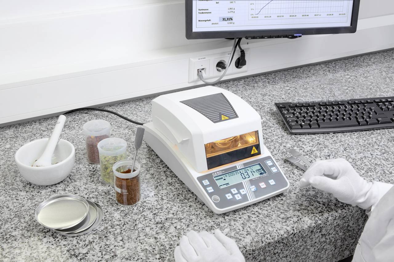 Ein Laborarbeitsplatz mit einem Feuchtemessgerät, umgeben von Probenbehältern und einem Computerbildschirm, auf dem Daten angezeigt werden.