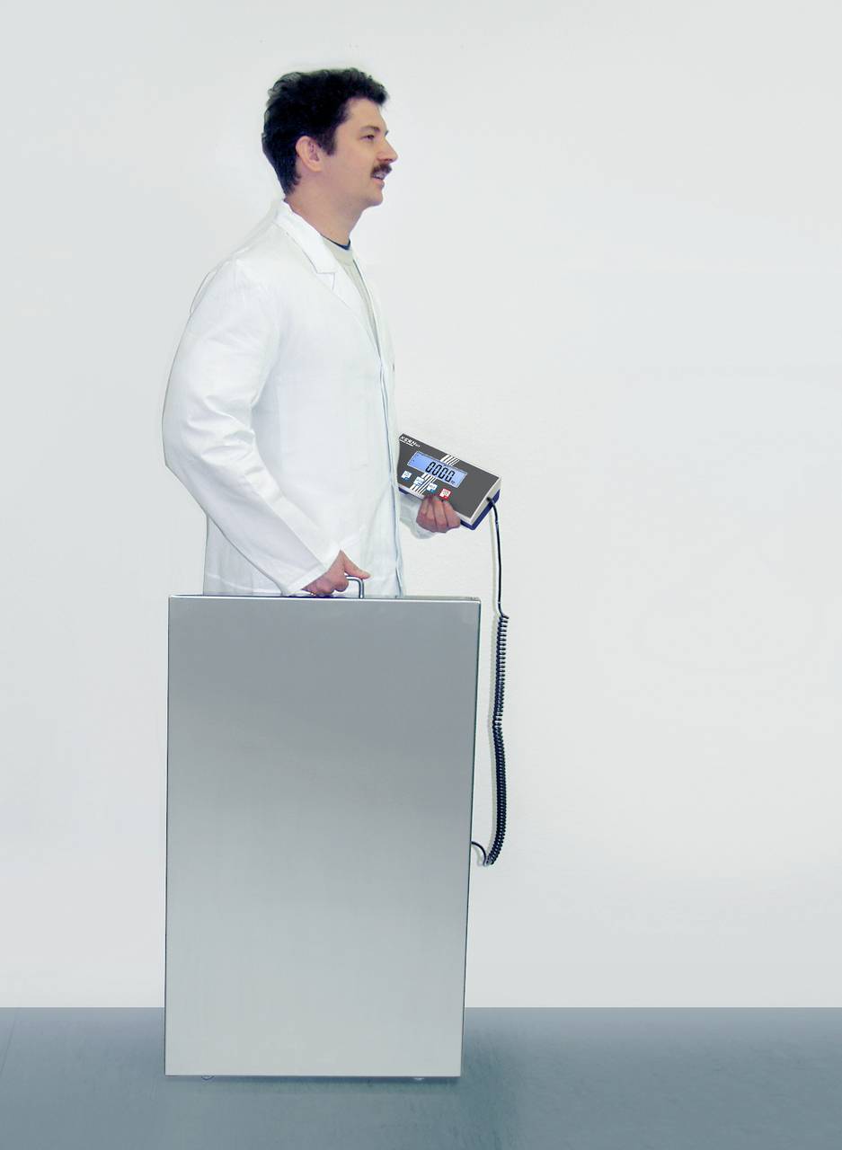 Ein Mann in weißem Kittel steht seitlich hinter einem großen, metallischen Gerät und hält ein Bedienfeld in der Hand.