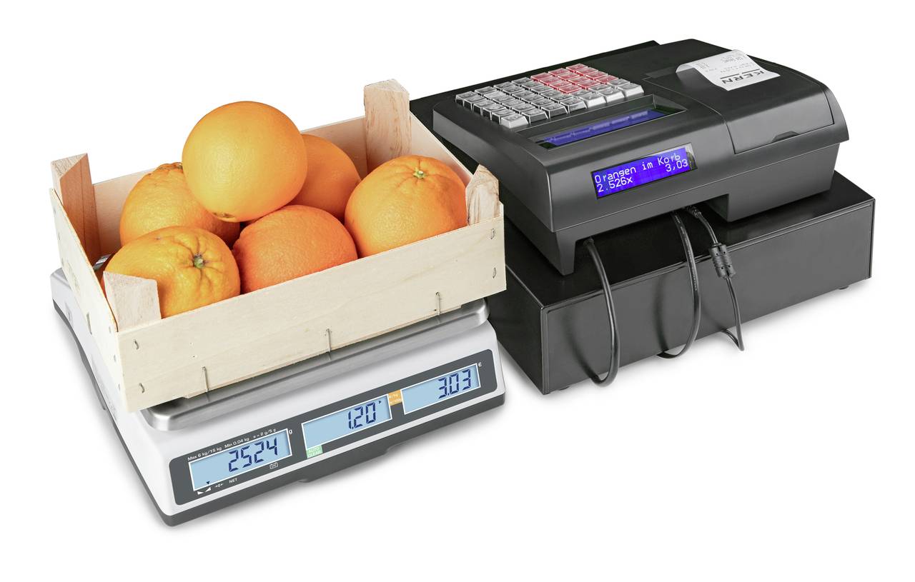 Eine Holzkiste mit Orangen steht auf einer digitalen Waage, die ein Gewicht von 2,524 kg anzeigt. Daneben steht eine elektronische Registrierkasse.